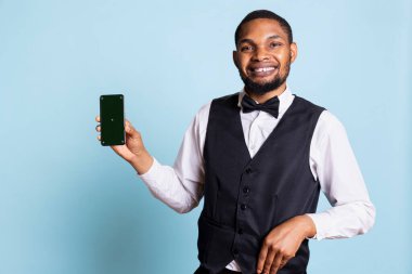 Otel çalışanı komi, stüdyoda mavi arka planda yeşil ekranı gösteren, izole edilmiş bir model ekranı olan telefona işaret ediyor. Otel kapıcısı resepsiyonda misafirleri karşılıyor..