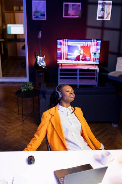 Oturma odasında ASMR 'ın rahatlatıcı seslerini dinleyen bir kadın, ses kulaklığı kullanarak, refahı artırmak için kendi kendine bakım yapıyor. Gözleri kapalı Afrikalı Amerikalı gece geç saatlerde evde ses terapisi yapıyor.