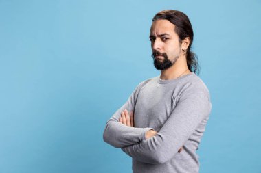 Mavi arka planda kendine güvenen bir erkek stüdyoda canlı ve havalı bir tavır sergiliyor. Kıvırcık uzun saçlı ve sakallı genç bir yetişkin kamerada kollarını kavuşturmuş poz veriyor..