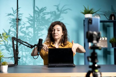 Stüdyodaki kadın, marka sponsoru videosundan dizüstü bilgisayarını onaylıyor, profesyonel mikrofonla konuşuyor. İçerik oluşturucu, yüksek teknoloji mikrofonuyla kayıt yaparak, dizüstü bilgisayarın tanıtımını yapıyor.