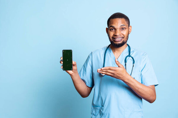 Портрет оптимистичного мужчины-медсестры, указывающего пальцем на зеленый экран на смартфоне. Уверенный работник больницы держит устройство с изолированным дисплеем хроматического ключа на фоне синей студии.