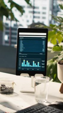 Aydınlık ofis ortamındaki cihazlarda analitik veri göstergelerine sahip dikey video Modern çalışma alanı. Finansal istatistik grafikleri saksı bitkilerinin yanında olan masadaki bilgisayar ekranı, dümen aşağı