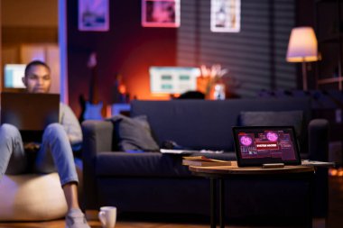 Siyahi bir adam bilgisayarında siber güvenlik için kodlama yaparken fasulye çuvalında oturuyor. Tablet, çevrimiçi güvenlik riskleri, veri hırsızlığı, kötü amaçlı yazılım ve yetkisiz erişim kaygılarını yansıtan bir siber saldırı konusunda uyarıyor.