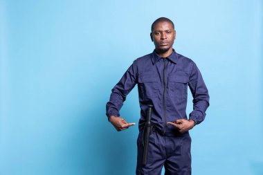 Stüdyoda kendi güvenliği için coplu silahını sunan silahlı bir güvenlik görevlisi düzen ve koruma sağlamak için polis yarasasını işaret ediyor. Mavi arka planda üniformalı erkek eğitimli personel.