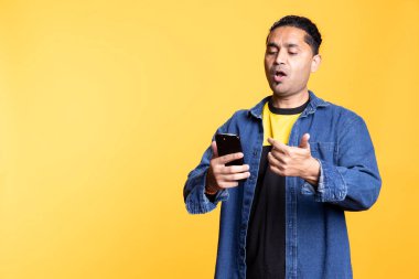 Erkek çöreği olan rahat Hintli adam akıllı telefon kullanıyor online uygulamaları tarıyor, sakince gülümsüyor ve turuncu bir stüdyonun arka planında duruyor. Modern teknolojiden ve kablosuz bağlantıdan hoşlanan adam.