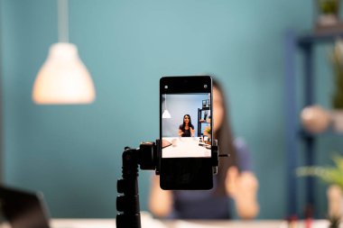 Akıllı telefon kamerası kozmetik ürünlerini inceleyen bir güzellik uzmanı yakalar. Mobil cihaz ekranında Asyalı vlogger 'ın mavi arka plan stüdyosunda güzellik ürünlerini açıklarken anlamlı jestler kullandığını gösteriyor..