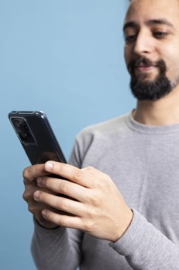 Dijital iletişimle uğraşırken akıllı telefonuyla çalışan düşünceli bir adam mavi arka planda duruyor. Modern çevrimiçi platformlarda uzaktan kumandalı iş yapan genç bir yetişkin..