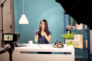 Neşeli kozmetik içeriği yaratıcısı, stüdyo ışıklandırmasıyla profesyonel bir düzenek kullanarak ruj markası değerlendirmesi yapıyor. Dijital reklamcılık, pazarlama ve sosyal medya eğilimleri için ideal.