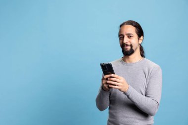 Modern dijital profesyonelliği somutlaştırmak için telefonu dalgın bir ifadeyle kullanan gündelik kıyafetlerle profesyonel bir genç adam. Havalı erkek modeli mavi arkaplanda aygıt kullanır.