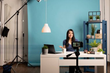 Kadın içerik yaratıcısı profesyonel bir stüdyoda makyaj dersi çekiyor. Neşeli Asyalı blogcu kozmetik ürünleriyle bir masada oturuyor, kameraya konuşuyor, ışıklandırıyor ve tripod kuruyor..