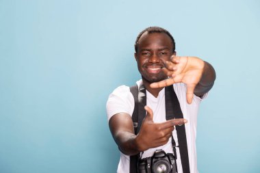 Afro-Amerikalı erkek fotoğrafçı kameraya bakmadan çekimini çerçeveliyor. Boynunda DSR ile parmaklarıyla dikdörtgen şekiller oluşturan siyah bir film yapımcısının portresi.