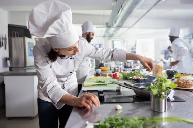 Modern mutfakta kadın aşçı ocağa lezzetli otlar ekliyor, taze yeşillik ve baharat kullanarak yemeği pişirirken lezzeti yükseltiyor. Lezzetli aşçılık sanatları, şef hayatı.