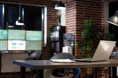 Modern ofiste boş bir masa, dizüstü bilgisayarı ve panosu olan, geceleri duvara yaslanmış. Arka plandaki bulanık bilgisayar monitörleri profesyonel ve minimalist bir çalışma alanı oluşturur.