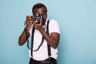 Beyaz tişörtlü kendinden emin bir erkek fotoğrafçı ve kamerasını tutan askılar fotoğrafçılığa olan tutkusunu gösteriyor. Stüdyo ayarları onun profesyonel ve şık görünümünü geliştirir..