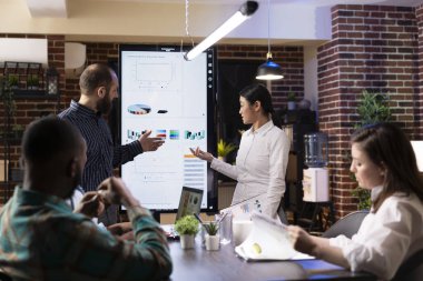 Asyalı ve Kafkasyalı iş arkadaşları, bir LED ekranının yanında duruyor, başlangıç ofisindeki beyin fırtınası sırasında finansal şemalar ve pazarlama stratejileri sunuyorlar. Takım marka stratejisinde işbirliği yapıyor..