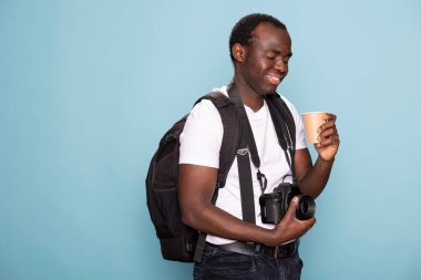 Afrikalı Amerikalı adam elindeki tek kullanımlık bardağa bakarken gülümseyerek kamera lensine dokunuyor. Sırt çantalı siyah erkek fotoğrafçı ve DSLR içerik ifadesiyle kahvesine bakıyor..
