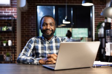 Modern bir ofiste masa başında oturan, tuğla duvarlı, kameraya bakan ve gülümseyen Afrikalı Amerikalı profesyonel bir erkek. Bilgisayarı ve panosu mali verileri incelemek için kurulmuş..