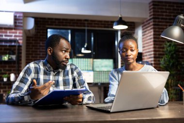 İki siyah profesyonel, bir dizüstü bilgisayar ve not panosundaki ön-market eğilimlerini gözden geçiriyorlar. Tuğla duvar ofisi, yoğun mali analiz ve takım çalışmasını vurguluyor.