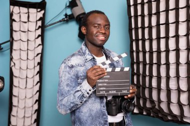 Afro-Amerikan erkek yönetmen profesyonel ışıklandırması olan bir stüdyoda, elinde bir klaket ve DSLR ile güven içinde poz veriyor. Elinde film tahtası olan siyah fotoğrafçı kameraya gülümsüyor..