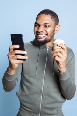 Afro-amerikan adam mesajlarını kontrol ediyor, kahve servisi yapıyor, kafein içkisini içiyor ve web sitelerini karıştırıyor. İnternette gezinen ve mesajlaşan genç kişi.