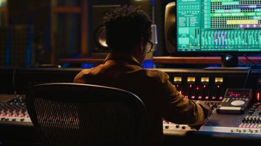 Afrika kökenli Amerikalı müzik yapımcısı profesyonel stüdyodaki yeni şarkısında kontrol odasında müzik karıştırıyor ve ustalaştırıyor. Teknisyen ses panelindeki düğmelere ve sürgülere basıyor. Kamera A.