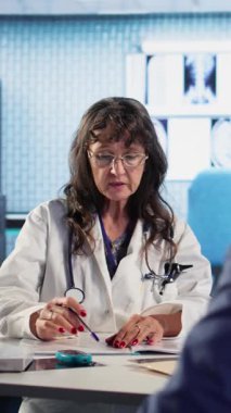 Dikey Videocu Kadın, rutin kontroller için bir erkek hastayı muayene ediyor. Klinikteki tedavi planı hakkında tartışıyorlar. Uzman, sigorta konusunda sağlık hizmetleri sunar. Kamera.