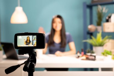 Tripoda monte edilmiş bir kameraya seçici bir odak noktası. Güzellik blogcusu kaydediyor. Cihaz ekranında gülümseyen Asyalı bir kadın makyaj dersleri ve eleştiriler üzerine canlı yayına başlarken el sallıyor..