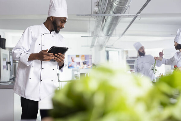 In a restaurant kitchen, african american chef in uniform holds laptop researching recipes while doing food prep with vegetables and other ingredients. Cooking gourmet dishes on the stove.