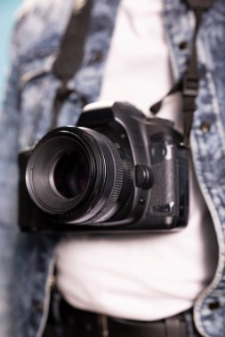 Siyah bir kameraya aşırı yakın çekim. Kot ceketli bir kişinin boynundan sarkan DSLR üzerine seçici bir odak noktası. Lense özel olarak odaklanmış. Karmaşık ayrıntıları vurguluyor..