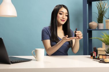 Genç güzellik blogcusu bir masada oturuyor, elinde ruj tutuyor ve internetteki izleyicilerine sunuyor. Canlı yayın seansı için bir makyaj videosu kaydederken ürüne doğru jest yapıyor..