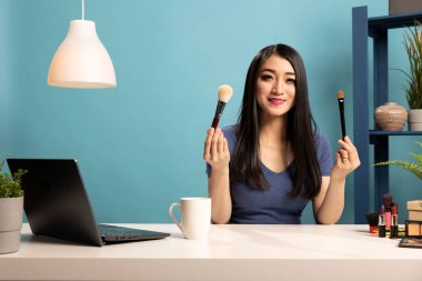 Gülümseyen güzel vlogger hakkında yaşayan kozmetik araçlar olmalıdır. Güzel Asyalı kadın masasında oturuyor, elinde makyaj fırçaları tutuyor, çevrimiçi izleyicileri için bir ürün değerlendirme videosu kaydediyor..