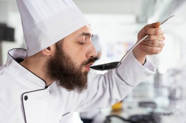 Mutfak mükemmelliğine odaklanan üniformalı erkek aşçı lezzetli bir çorbanın tadına bakmak için kaşık kullanıyor, çeşnisini gurme yemeğine göre ayarlıyor. Mutfak aromatik malzemelerle dolu. Kapat..