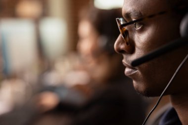 Gözlük takan siyahi bir adamın internet destek ofisindeki müşterilerle iletişim kurması çok yakın bir plan. Afrika kökenli Amerikalı erkek ajan, kulaklık takıyor, müşterilere yardım ediyor..