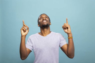 Neşeli Afro-Amerikalı adam yukarıya bakıyor, sanki bir mesaj veriyor gibi. Mavi arka planda rahat bir tişört giyen mutlu bir erkek. Reklam için mükemmel..