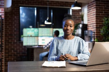 Modern ofiste gece geç saatlere kadar çalışan siyah bir kadın masasında dizüstü bilgisayarını ve panosunu kullanıyor. Afrika kökenli Amerikalı kadın borsa analisti masada oturmuş kameraya bakıyor ve vardiyasına hazırlanıyor..