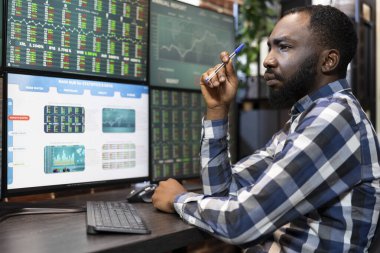 Masada geç saatlere kadar çalışan odaklanmış bir siyah adam modern bir ofisteki birden fazla monitördeki borsa verilerini analiz ediyor. Ticaret istatistiklerini incelerken elinde kalem tutan borsacı..