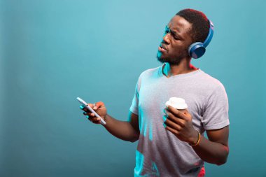 Mavi kulaklık takan, elinde kahve tutan ve telefonuna şok edici bir ifadeyle bakan bir adam. Tek kullanımlık bardağı olan Afro-Amerikalı adam cep telefonundan gelen şok edici haberlere şaşırdı..