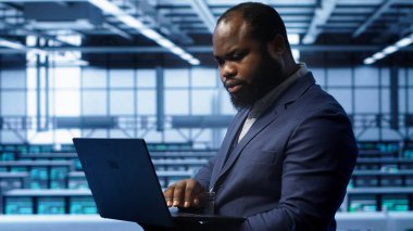 Sunucu merkezindeki yazılım geliştiricisi dizüstü bilgisayardaki kurtarma planını kontrol ediyor, donanım enerji tüketimini izliyor. Afrikalı Amerikalı işçiler veri merkezinde dizüstü bilgisayar kullanıyor, teçhizatların çalışmasını sağlıyorlar, kamera A