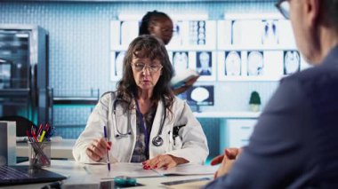 Kıdemli doktor ve hasta modern bir tıp ofisinde teşhis sonuçlarını tartışıyor, gelişmiş teknolojiyi röntgen ve MRI taramalarını gösteren kayıtlar kullanarak sergiliyorlar. Bakım yönetimi. Kamera A.