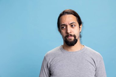 Mavi arka planda kendine güvenen bir erkek stüdyoda canlı ve havalı bir tavır sergiliyor. Kıvırcık uzun saçlı ve sakallı genç bir yetişkin kamerada kollarını kavuşturmuş poz veriyor..