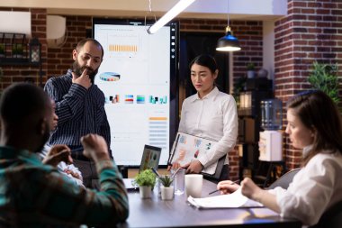 İş adamları pazarlama stratejilerini LED ekrandaki dijital çizelgeleri kullanarak gözden geçiriyorlar. Çeşitli meslektaşlar, gece geç saatlerde yapılan toplantıda finansal analiz ve çevrimiçi veri içgörüleri için modern teknolojiyi kullandılar..