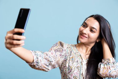 Sosyal medyada selfie çekmek için akıllı telefonunu kullanan mutlu bir insan. Neşeli Hintli kadın fotoğraf çekiyor. Telefon kamerasıyla. Stüdyonun mavi arka planında duruyor..