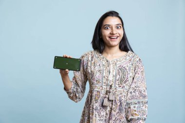 Mobil cihazda yeşil ekran sunan kendine güvenen bir kadın stüdyo arka planına karşı duruyor. Neşeli Hintli kişi, reklam markalarını kopyalamak için kullanılan krom anahtarlı akıllı telefonu tutuyor..