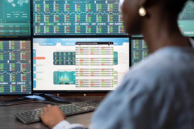 Ofis masasındaki klavyeye yazı yazan siyahi kadın çalışanın yakın çekimi, birden fazla ekranda borsa ilerlemesini gözden geçiriyor. Ticari istatistikleri ve verileri gösteren dijital monitöre seçici odak.