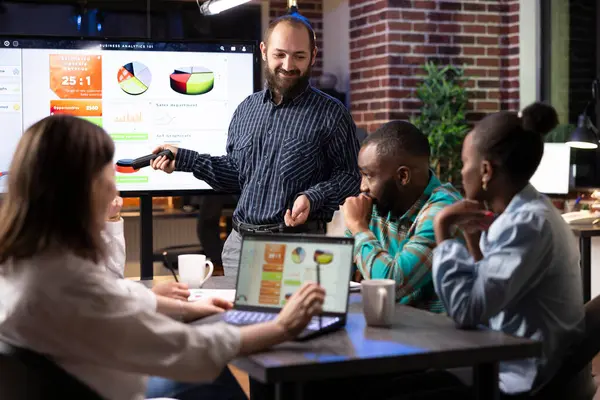 Kafkasyalı bir adam, modern çalışma ortamındaki gece toplantısında iş arkadaşları mali verileri analiz ederken iş sunumu yapıyor. Stratejik planlama oturumunu dizüstü bilgisayar, belgeler ve takım çalışması yürütüyor.