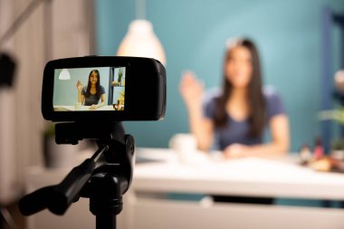 Canlı yayını için güzellik etkilerini kaydeden tripodun üzerindeki dijital kamera. Cihaz ekranı masasında oturan gülümseyen Asyalı bir kadına odaklanıyor. Stüdyoda vlog yaparken el sallıyor..