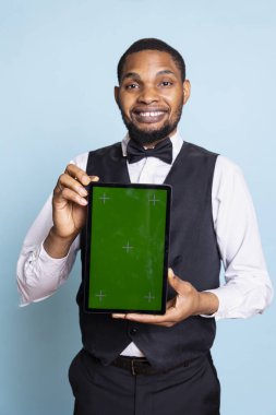Stüdyoda krom ekranlı tablet tutan gülümseyen bir otelci profesyonel smokin üniforması giyiyor ve müşterileri selamlıyor. Mavi arka planda genç otel kapıcısı..