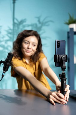 Stüdyoda izleyicilerle tartışmak için neşeli bir internet yıldızının kullandığı selfie çubuğuna bağlı akıllı telefona odaklanın. Neşeli kadının sosyal medya için video kaydetmek için kullandığı telefonu kapat