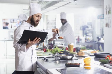 Mutfak araştırmalarına odaklanan, yoğun bir restoran mutfağındaki erkek aşçı, malzemeleri hazırlarken yeni tarif fikirleri bulmak için dizüstü bilgisayar kullanıyor. Adam gurme yemekleri pişiriyor ve lezzetli yemekler hazırlıyor..