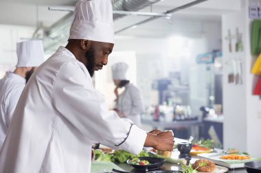 Afro-amerikan şef biberiyeli ya da fesleğenli lezzetli yemekler pişiriyor restoranın mutfağında, fırında bitki yaprakları serpiyor. Genç aşçı taze ürün ve hammadde kullanıyor..
